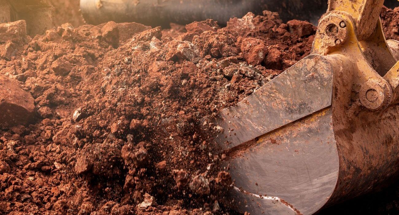 Nahaufnahme einer Baggerschaufel, die sich durch rotbraune Erde gräbt und dabei Staub und Schmutz aufwirbelt.